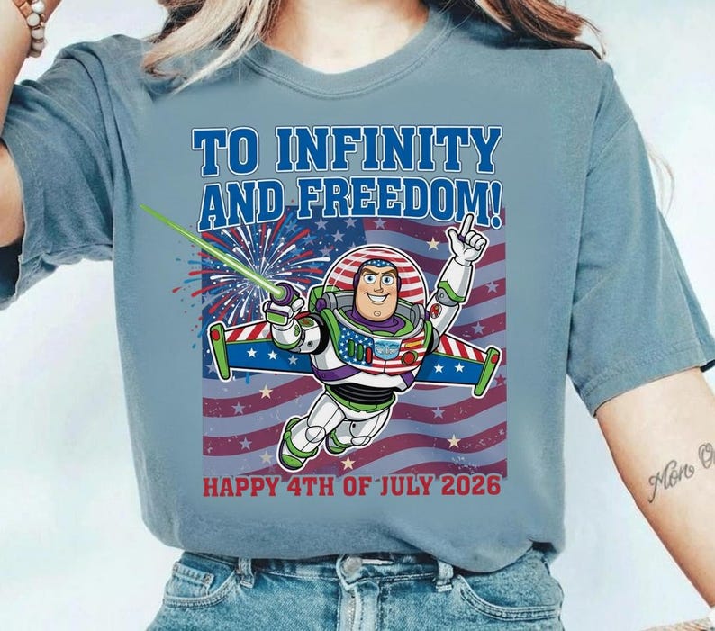 May include: Light blue t-shirt featuring Buzz Lightyear with the text "TO INFINITY AND FREEDOM!" and "HAPPY 4TH OF JULY 2026" over an American flag design. Buzz is holding a green laser and pointing upwards.
