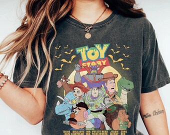 May include: Dark gray t-shirt featuring the words "TOY STORY" in green and yellow, with a graphic of characters from the movie. The characters include Woody, Buzz Lightyear, and Rex. The shirt also has the names of the characters at the bottom.