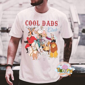 May include: White t-shirt with the text "COOL DADS Club" in red and blue. The shirt features a colorful cartoon graphic of various animated characters. The shirt is made of a soft material.