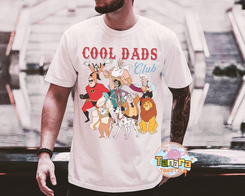 May include: White t-shirt with the text "COOL DADS Club" in red and blue. The shirt features a colorful cartoon graphic of various animated characters. The shirt is made of a soft material.