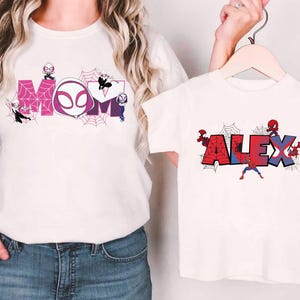 Puede incluir: Camisetas blancas con dise&ntilde;os de superh&eacute;roes. Una camiseta presenta la palabra "MOM" en rosa y morado con detalles de telara&ntilde;as y personajes de superh&eacute;roes. La otra camiseta muestra el nombre "ALEX" en rojo y azul con detalles de telara&ntilde;as y personajes de superh&eacute;roes.