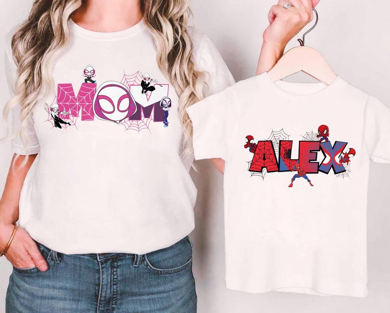 Puede incluir: Camisetas blancas con dise&ntilde;os de superh&eacute;roes. Una camiseta presenta la palabra "MOM" en rosa y morado con detalles de telara&ntilde;as y personajes de superh&eacute;roes. La otra camiseta muestra el nombre "ALEX" en rojo y azul con detalles de telara&ntilde;as y personajes de superh&eacute;roes.