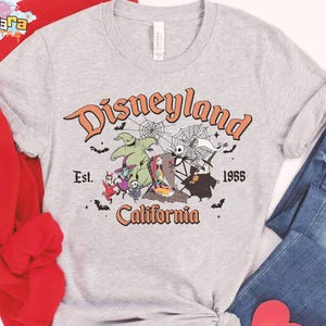 May include: A heather gray t-shirt featuring a graphic print with the text Disneyland California Est. 1955. The design includes characters from The Nightmare Before Christmas, spiderwebs, and bats in orange, black, and white colors on the front.
