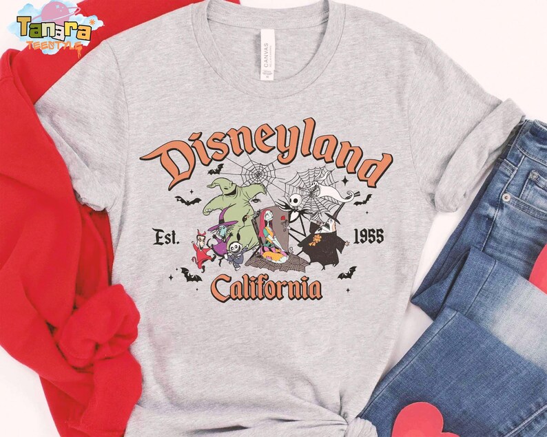 May include: A heather gray t-shirt featuring a graphic print with the text Disneyland California Est. 1955. The design includes characters from The Nightmare Before Christmas, spiderwebs, and bats in orange, black, and white colors on the front.