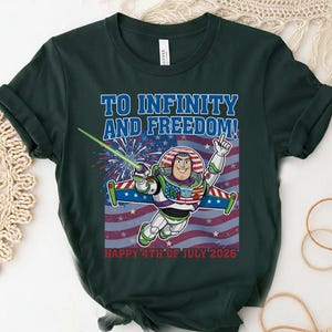 May include: Dark green t-shirt featuring a graphic of Buzz Lightyear in a spacesuit with the text "TO INFINITY AND FREEDOM!" and "HAPPY 4TH OF JULY 2026" against a red, white, and blue American flag background.
