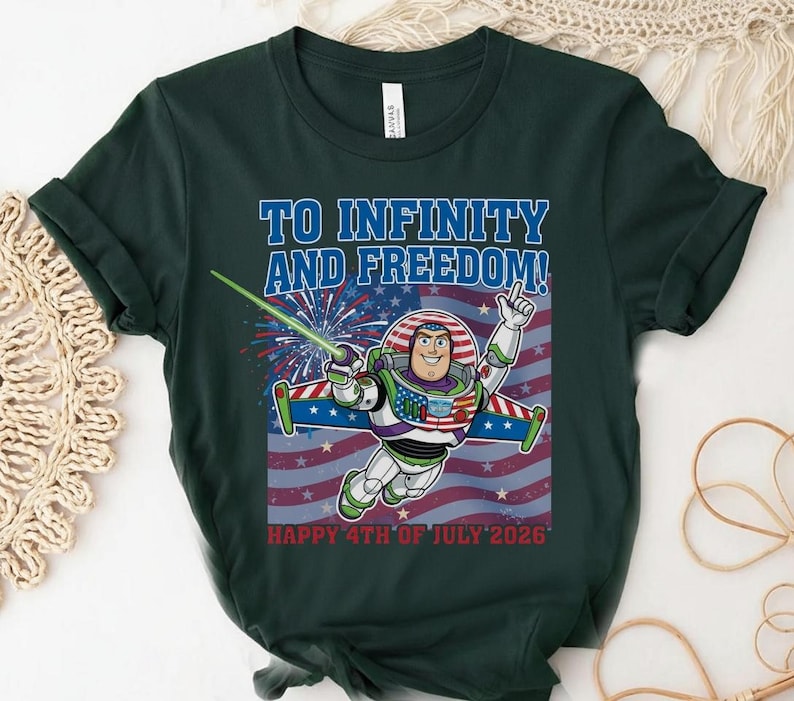 May include: Dark green t-shirt featuring a graphic of Buzz Lightyear in a spacesuit with the text "TO INFINITY AND FREEDOM!" and "HAPPY 4TH OF JULY 2026" against a red, white, and blue American flag background.