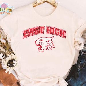 May include: Cream-colored t-shirt with the words "EAST HIGH" in red block letters above a red and white wildcat graphic. The shirt is a casual style, suitable for everyday wear.