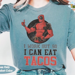 May include: A blue jean-colored t-shirt with a graphic of Deadpool giving a thumbs up. The text reads "I WORK OUT SO I CAN EAT TACOS" in a distressed font. The shirt is a comfort color.