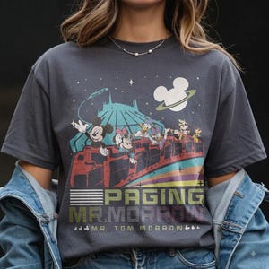 May include: A charcoal gray t-shirt featuring a graphic of Mickey Mouse, Minnie Mouse, Donald Duck, and Daisy Duck riding a space-themed roller coaster. The design includes a Mickey-shaped planet and the text PAGING MR. MORROW MR. TOM MORROW.