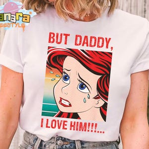 May include: White t-shirt featuring a distressed pop art style illustration of a cartoon character with red hair and blue eyes, with the text "BUT DADDY, I LOVE HIM!!!" in red. The shirt has a crew neck and short sleeves.