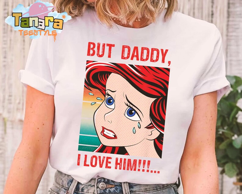 May include: White t-shirt featuring a distressed pop art style illustration of a cartoon character with red hair and blue eyes, with the text "BUT DADDY, I LOVE HIM!!!" in red. The shirt has a crew neck and short sleeves.
