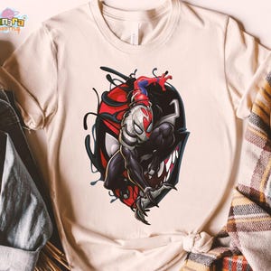 May include: Cream-colored t-shirt featuring a graphic of a superhero character in a black and red design. The character is depicted in a dynamic pose, with a detailed illustration style. The shirt has a classic crew neck and short sleeves.