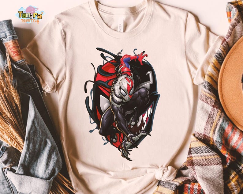 May include: Cream-colored t-shirt featuring a graphic of a superhero character in a black and red design. The character is depicted in a dynamic pose, with a detailed illustration style. The shirt has a classic crew neck and short sleeves.
