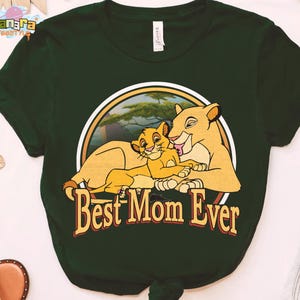 May include: Dark green t-shirt featuring a circular graphic with a lioness and cub, with the text "Best Mom Ever" in a stylized font. The graphic is set against a background of a savanna landscape.