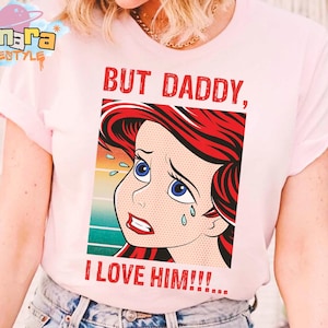 May include: Light pink t-shirt featuring a pop art style illustration of a crying cartoon character with red hair and blue eyes. The text "BUT DADDY, I LOVE HIM!!!" is printed in red. The shirt has short sleeves.