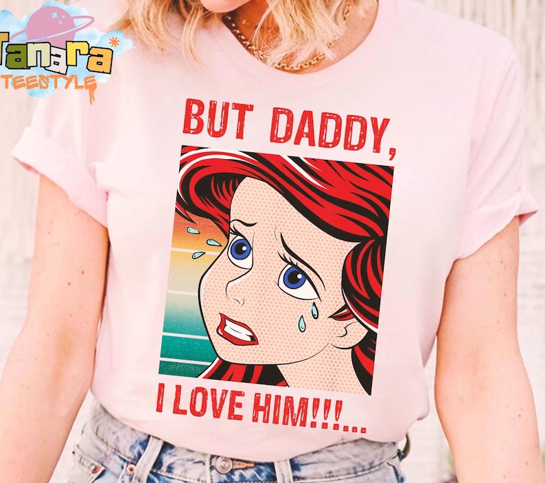May include: Light pink t-shirt featuring a pop art style illustration of a crying cartoon character with red hair and blue eyes. The text "BUT DADDY, I LOVE HIM!!!" is printed in red. The shirt has short sleeves.