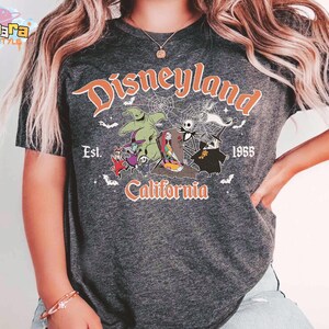 May include: A dark heather gray t-shirt featuring a graphic print with the text Disneyland Est. 1955 California. The design includes characters from The Nightmare Before Christmas, spiderwebs, and bats in orange, white, and green colors.