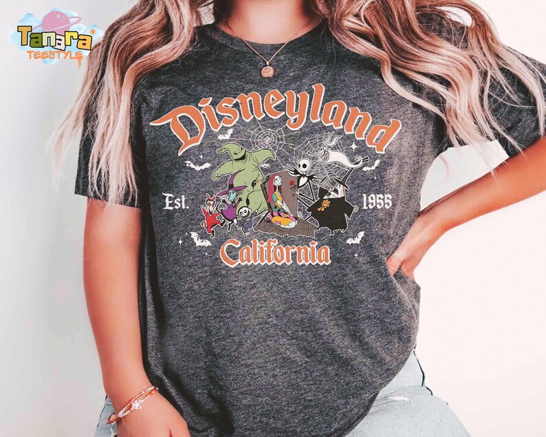 May include: A dark heather gray t-shirt featuring a graphic print with the text Disneyland Est. 1955 California. The design includes characters from The Nightmare Before Christmas, spiderwebs, and bats in orange, white, and green colors.