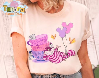 Puede incluir: Camiseta color crema con una ilustración caprichosa del Gato de Cheshire de Alicia en el país de las maravillas. El diseño incluye tazas apiladas, globos y mariposas. El texto "Tanara TEENSTYLE" está impreso en la parte superior izquierda.