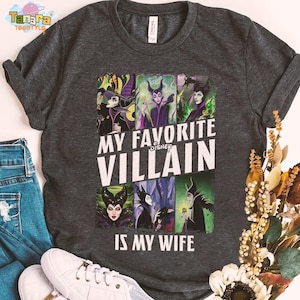 May include: Dark gray t-shirt featuring a collage of Disney villains with the text "MY FAVORITE VILLAIN IS MY WIFE". The shirt is paired with blue jeans and white sneakers. The shirt is made of a soft material.