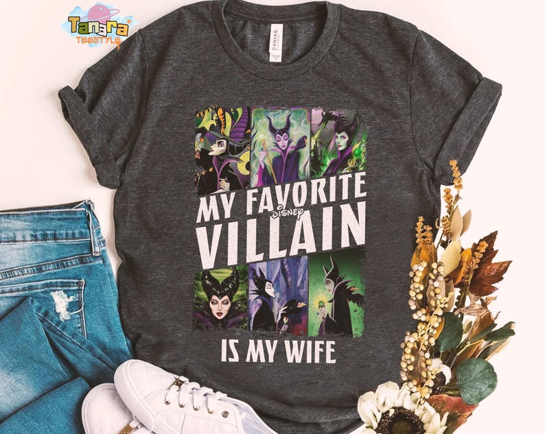 May include: Dark gray t-shirt featuring a collage of Disney villains with the text "MY FAVORITE VILLAIN IS MY WIFE". The shirt is paired with blue jeans and white sneakers. The shirt is made of a soft material.