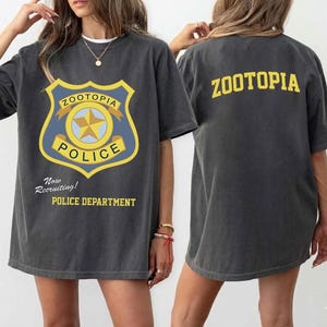May include: Dark gray t-shirt featuring a Zootopia Police Department badge graphic on the front and the word "ZOOTOPIA" on the back. The front graphic includes the text "Now Recruiting!" and "POLICE DEPARTMENT".