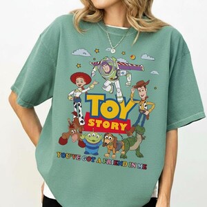 May include: A sage green t-shirt featuring a colorful graphic of characters from the animated film "Toy Story." The design includes Woody, Buzz Lightyear, Jessie, and other characters, with the words "Toy Story" and "You've Got a Friend in Me."