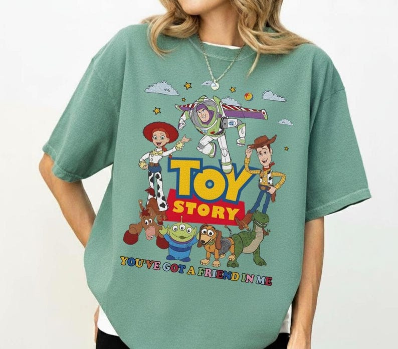 May include: A sage green t-shirt featuring a colorful graphic of characters from the animated film "Toy Story." The design includes Woody, Buzz Lightyear, Jessie, and other characters, with the words "Toy Story" and "You've Got a Friend in Me."
