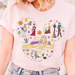 May include: Light pink t-shirt featuring a heart-shaped design with illustrations of characters and elements from the animated movie "Tangled." The design includes Rapunzel, a horse, and floating lanterns. The shirt has the text "Tanara TeeStyle" at the top.