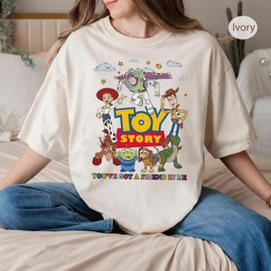 May include: Ivory-colored t-shirt featuring a colorful graphic of Toy Story characters, including Woody, Buzz Lightyear, and Jessie. The words "TOY STORY" are in large, bold letters, with the phrase "YOU'VE GOT A FRIEND IN ME" below.