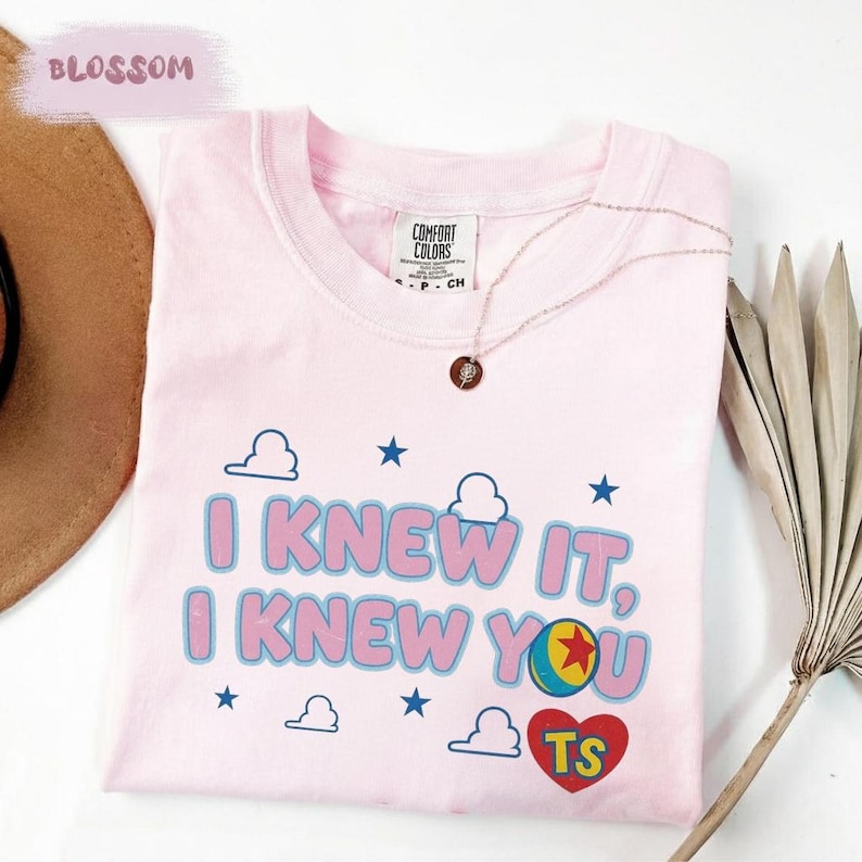 K&ouml;nnte beinhalten: Ein hellrosa T-Shirt mit kurzen &Auml;rmeln und dem Text I KNEW IT, I KNEW YOU in blauen und rosa Buchstaben. Das Design enth&auml;lt blaue Sterne, wei&szlig;e Wolken, einen bunten Ball mit einem roten Stern und ein rotes Herz mit den gelben Buchstaben TS.