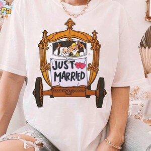 May include: White t-shirt featuring a cartoon carriage with a bride and groom, and a fox. The carriage has a gold frame and brown wheels. The sign on the carriage reads "JUST MARRIED" with two pink hearts.