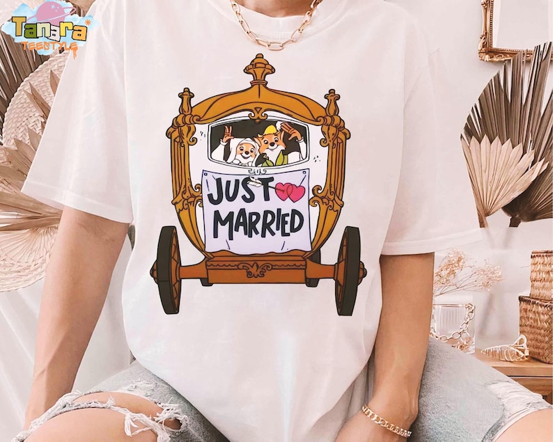 May include: White t-shirt featuring a cartoon carriage with a bride and groom, and a fox. The carriage has a gold frame and brown wheels. The sign on the carriage reads "JUST MARRIED" with two pink hearts.