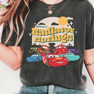 Pode incluir: T-shirt cinzento escuro com um gr&aacute;fico de personagens do filme Carros, incluindo o Rel&acirc;mpago McQueen. O design inclui o texto "Welcome to Radiator Springs" em amarelo e verde, com um sol e nuvens retr&ocirc;.
