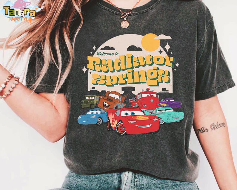 Pode incluir: T-shirt cinzento escuro com um gr&aacute;fico de personagens do filme Carros, incluindo o Rel&acirc;mpago McQueen. O design inclui o texto "Welcome to Radiator Springs" em amarelo e verde, com um sol e nuvens retr&ocirc;.