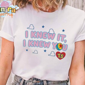 K&ouml;nnte beinhalten: Ein wei&szlig;es Kurzarm-T-Shirt mit dem rosa Schriftzug I KNEW IT, I KNEW YOU in blauer Umrandung. Das Design enth&auml;lt kleine blaue Sterne, wei&szlig;e Wolkenumrisse, einen bunten Ball mit einem roten Stern und ein rotes Herz mit den Buchstaben TS.