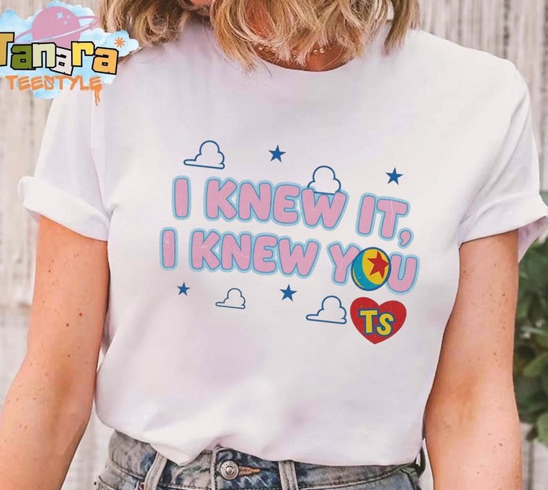 K&ouml;nnte beinhalten: Ein wei&szlig;es Kurzarm-T-Shirt mit dem rosa Schriftzug I KNEW IT, I KNEW YOU in blauer Umrandung. Das Design enth&auml;lt kleine blaue Sterne, wei&szlig;e Wolkenumrisse, einen bunten Ball mit einem roten Stern und ein rotes Herz mit den Buchstaben TS.