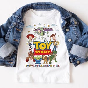 May include: White t-shirt featuring the Toy Story characters Buzz Lightyear, Woody, Jessie, and others. The shirt has the words "TOY STORY" in yellow and red, with the phrase "YOU'VE GOT A FRIEND IN ME" below. A blue denim jacket is draped over the shirt.