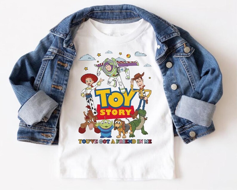 May include: White t-shirt featuring the Toy Story characters Buzz Lightyear, Woody, Jessie, and others. The shirt has the words "TOY STORY" in yellow and red, with the phrase "YOU'VE GOT A FRIEND IN ME" below. A blue denim jacket is draped over the shirt.
