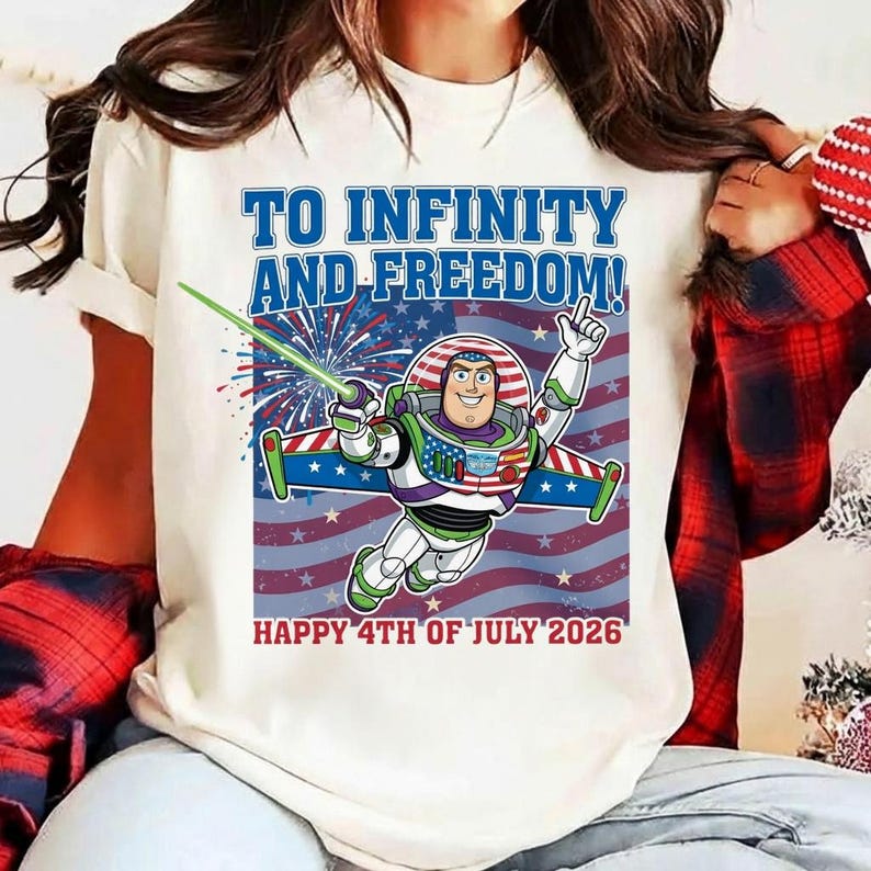 May include: Cream-colored t-shirt featuring a graphic of Buzz Lightyear with the text "TO INFINITY AND FREEDOM!" and "HAPPY 4TH OF JULY 2026". The design includes a red, white, and blue American flag background and fireworks.