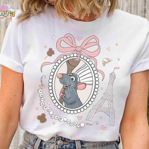 May include: White t-shirt featuring a cartoon rat holding a whisk inside an oval frame, with a pink bow, Eiffel Tower, and the text "ANYONE CAN COOK". The design includes hearts and pastries.