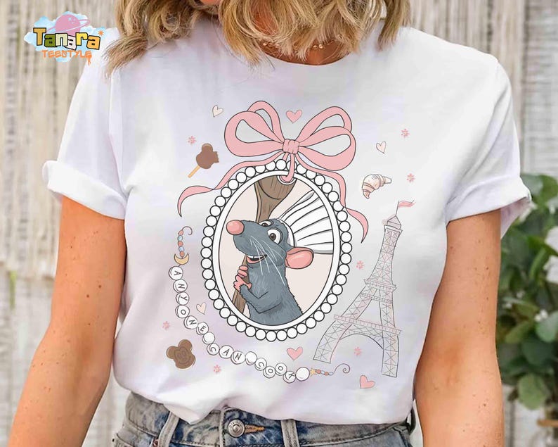May include: White t-shirt featuring a cartoon rat holding a whisk inside an oval frame, with a pink bow, Eiffel Tower, and the text "ANYONE CAN COOK". The design includes hearts and pastries.