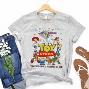 Puede incluir: Camiseta gris claro con los personajes de Toy Story: Buzz Lightyear, Woody y Jessie. La camiseta tiene las palabras "TOY STORY" y "YOU'VE GOT A FRIEND IN ME" impresas. Tambi&eacute;n hay vaqueros azules y sandalias en la imagen.
