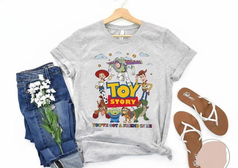 Puede incluir: Camiseta gris claro con los personajes de Toy Story: Buzz Lightyear, Woody y Jessie. La camiseta tiene las palabras "TOY STORY" y "YOU'VE GOT A FRIEND IN ME" impresas. Tambi&eacute;n hay vaqueros azules y sandalias en la imagen.