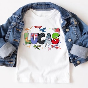 Op de afbeelding: Wit T-shirt met de naam "LUCAS" in kleurrijke, gestileerde letters, elk met een uniek ontwerp. Het shirt heeft verschillende cartoonvliegtuigen en het woord "PLANES". Een spijkerjasje is over het shirt gedrapeerd.