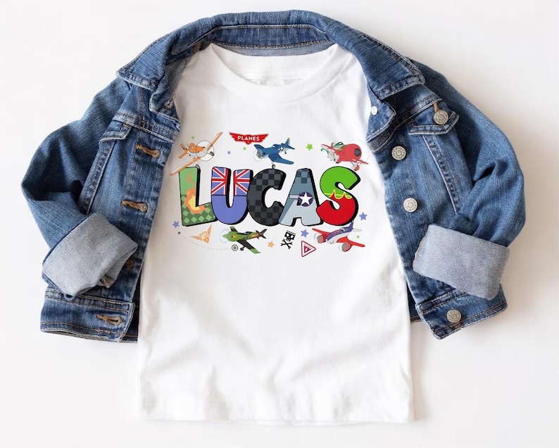 Op de afbeelding: Wit T-shirt met de naam "LUCAS" in kleurrijke, gestileerde letters, elk met een uniek ontwerp. Het shirt heeft verschillende cartoonvliegtuigen en het woord "PLANES". Een spijkerjasje is over het shirt gedrapeerd.