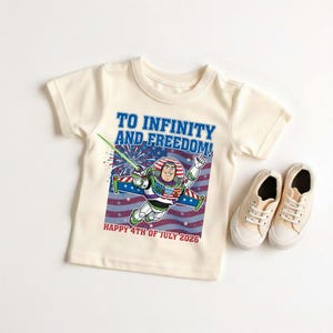 May include: Cream-colored t-shirt featuring a graphic of Buzz Lightyear in a patriotic design with the text "TO INFINITY AND FREEDOM!" and "HAPPY 4TH OF JULY 2026". A pair of beige canvas sneakers are next to the shirt.