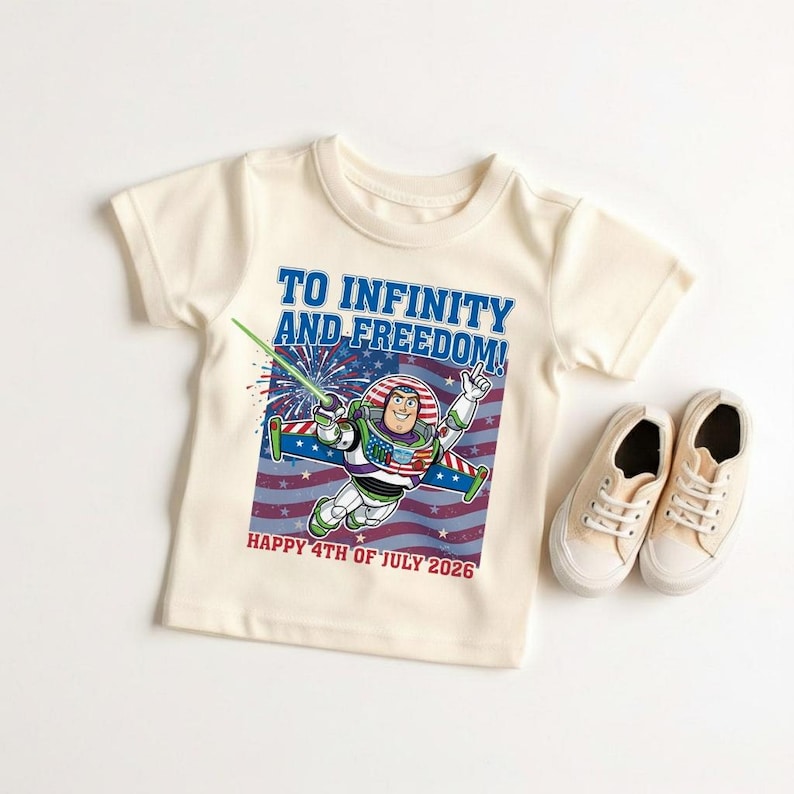 May include: Cream-colored t-shirt featuring a graphic of Buzz Lightyear in a patriotic design with the text "TO INFINITY AND FREEDOM!" and "HAPPY 4TH OF JULY 2026". A pair of beige canvas sneakers are next to the shirt.