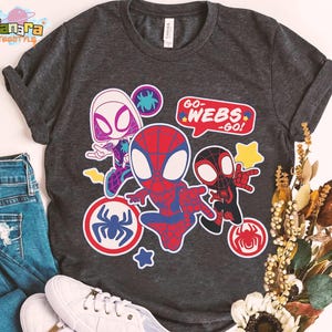 May include: Dark gray t-shirt featuring cartoon illustrations of Spider-Man characters. The design includes Spider-Gwen, Spider-Man, and Miles Morales, with the text "GO-WEBS-GO!" in a speech bubble. Additional spider web and star graphics are also present.