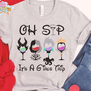 May include: Pale grey t-shirt with the words "OH SIP" and "It's A Girls Trip". The design shows four wine glasses with cartoon character-inspired designs, each filled with a different coloured liquid. The shirt is made of a soft material.